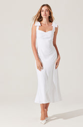 A woman is wearing a sleeveless white dress that has wide shoulder straps with prominent bow-like details on each shoulder, a fitted bodice with a sweetheart neckline, and a long, flowing skirt that reaches to her ankles. She is also wearing white pointed-toe shoes.